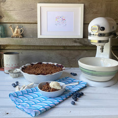 a blueberry crumble in a white pie dish, a small bowl of blueberry crumble with whipped cream, on a rustic white table with a blue fish scale patterned tea towel and stand mixer to the side