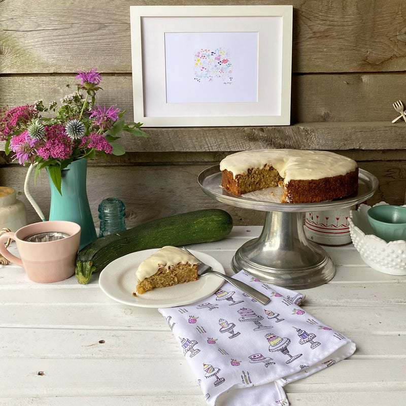 a slice of zucchini cake with cream cheese frosting on a plate on a rustic white table, the rest of the cake on a cake stand with fresh flowers in a vase, and a cute cotton tea towel with cakes and cupcakes on the table